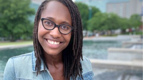 K. Tahji Claybren, a black, queer, non-binary person with dark skin and chin-length locs wearing black cat-eye glasses, a blue-jean jacket and blue shirt smiles in front of a public water feature.