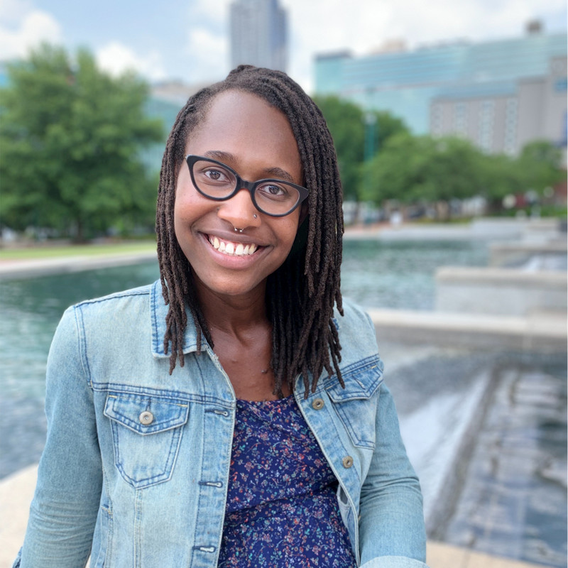 K. Tahji Claybren, a black, queer, non-binary person with dark skin and chin-length locs wearing black cat-eye glasses, a blue-jean jacket and blue shirt smiles in front of a public water feature.