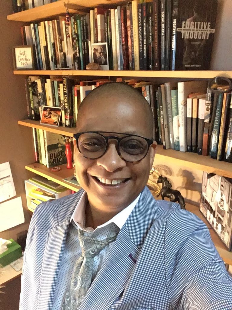 Dr. Sharon P. Holland smiles in front of a full bookshelf, wearing a gray suit and black glasses.
